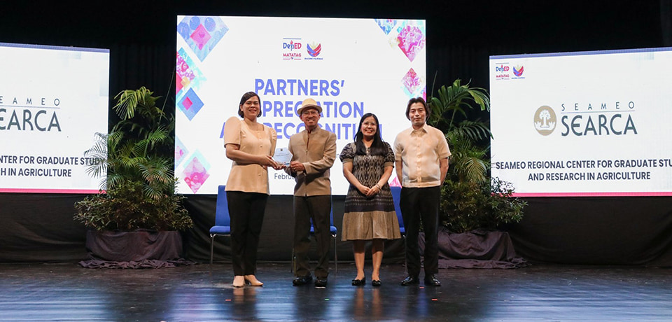 Vice President of the Republic of the Philippines and DepEd Secretary H.E. Sara Zimmerman Duterte (leftmost) hands over the appreciation plaque to Dr. Glenn Gregorio (second to the left), SEARCA Center director.  With them on the stage are Ms. Beatrisa Martinez (second to the right), executive coordinator of the Office of the Director, and Mr. Michael Poa (rightmost), DepEd spokesperson. [Photo by Alezandro Bravo, grabbed from the DepEd Philippines Facebook page]
